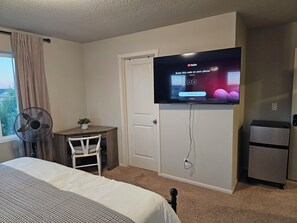 A hotel room with a bed, a desk with a chair, a TV mounted on the wall, a fan, and a refrigerator.