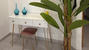 Een kamer met een wit bureau, twee blauwe vazen, een spiegel, een grote groene plant en een houten stoel.