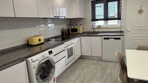 Een moderne keuken met witte kasten, een wasmachine, een magnetron, en een eethoek met een tafel en stoelen.