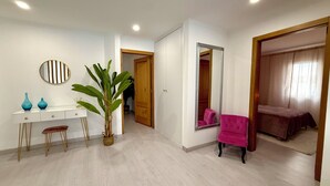 Een kamer met een bureau, twee stoelen, een grote spiegel, een kamerplant en een bed met een roze stoel.