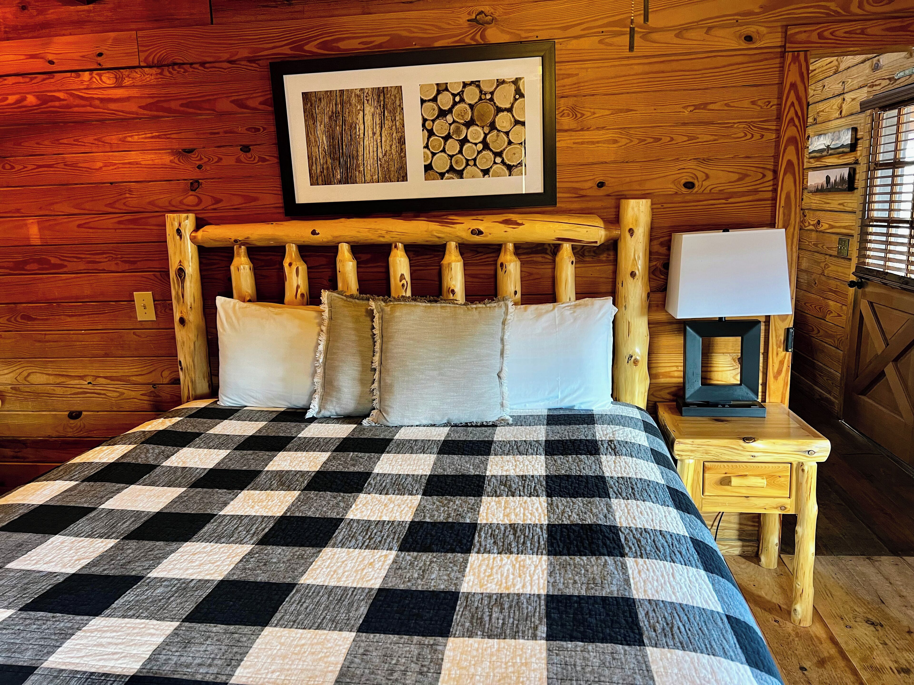 Interno di una baita di legno con una coperta a quadri, un quadro incorniciato sopra il letto e un comodino con una lampada.