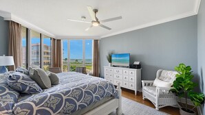 Une chambre avec un lit, une télévision, une commode blanche, un fauteuil en osier et une vue sur l’océan.