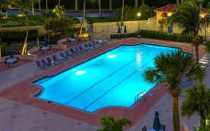Ein Pool mit Liegestühlen und Sonnenschirmen, umgeben von Palmen und einem Gebäude im Hintergrund.