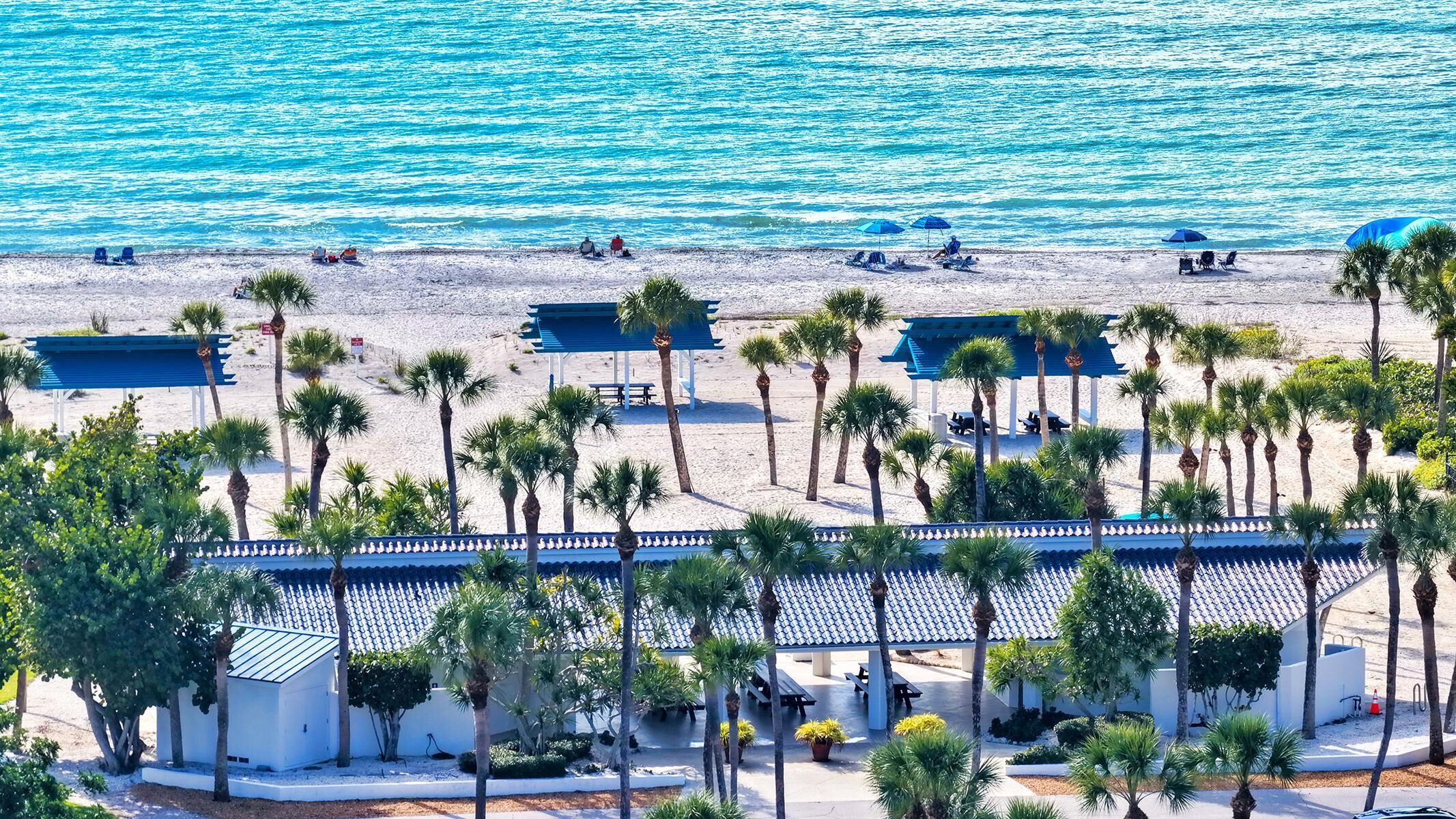 A beach with palm trees, beach umbrellas, and a covered area with benches.