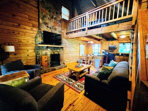 A cabin interior with a stone fireplace, wooden furniture, a balcony, and a kitchen area.