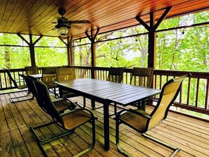 A covered outdoor dining area with a table and chairs, wooden flooring, and a view of trees through the railing. - Perfect cabin for large families (Warm Springs)
