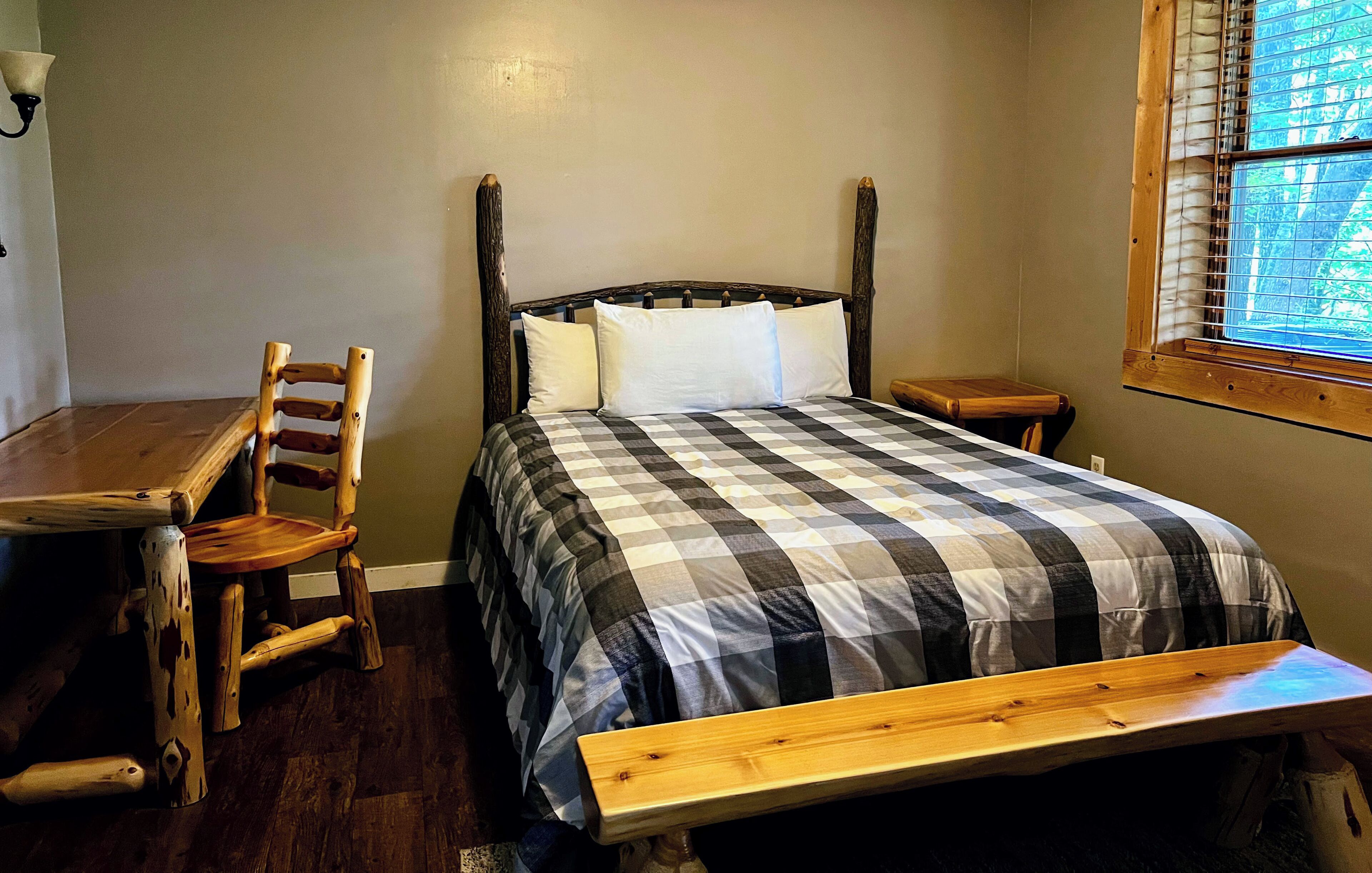 A bedroom with a wooden bed, a checkered bedspread, a wooden chair, a small wooden table, and a window with blinds.
