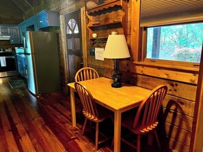 A wooden cabin interior with a dining table, chairs, a lamp, a refrigerator, and a window with a view of trees. - Beautiful sunsets, decks, firepit! (Warm Springs)