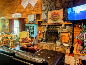 A cozy living room with a stone fireplace, a television, and a dining area with wooden furniture. - Shabby Chic meets the mountains! Sleeps 6 (Warm Springs)