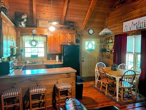 A wooden cabin interior with a kitchen, dining area, and a visible door with a wreath. - Shabby Chic meets the mountains! Sleeps 6 (Warm Springs)
