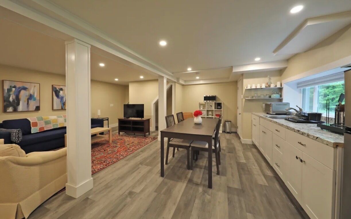 A modern living room with a sofa, a coffee table, and a TV. There is a dining area with a table and chairs. The kitchen has cabinets and a window.