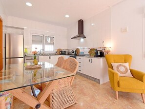 Una cocina moderna con armarios blancos, una mesa de comedor de vidrio, sillas de mimbre y un sillón amarillo.