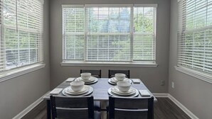 Ein Esszimmer mit einem Tisch für vier Personen, großen Fenstern mit Jalousien und einer Deckenleuchte.