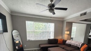 Ein Wohnzimmer mit Ecksofa, Deckenventilator, großem Fenster und an der Wand befestigtem Fernseher.