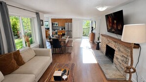 Sala de estar com sofá, mesa de centro, lareira, TV e vista para a cozinha e o jardim.