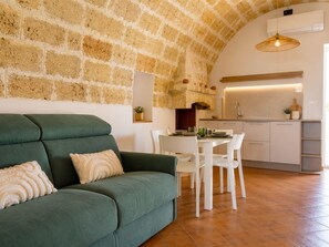 A cozy living area with a green sofa, white dining table, and chairs, a kitchen with a brick wall, and a hanging light fixture. - Tenuta Polignano - La Scuderia, a Rural Estate in Puglia.  Exposed Stone Arches (Puglia)