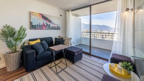 Uma sala de estar moderna com sofá, puf e uma mesa pequena. Há uma janela grande com vista para edifícios e montanhas.