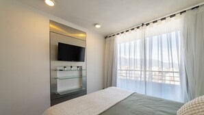 Quarto com cama, TV fixada na parede, estante de vidro e janela com cortinas leves.
