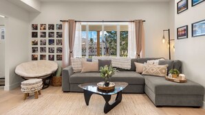 En moderne stue med en grå hjørnesofa, et glasbord og et stort vindue med gardiner.