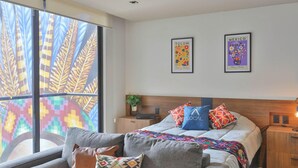 Ein Schlafzimmer mit einem Bett, Kissen, einem Nachttisch, einer Lampe und einem Fenster mit Blick auf farbenfrohe Muster.