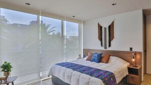 Um quarto moderno com uma cama grande, uma mesinha de cabeceira, uma luminária na parede e vista através de uma porta de vidro deslizante.