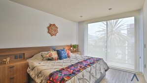 Quarto com cama, mesa de cabeceira, objeto decorativo na parede e vista para palmeiras através de uma porta de correr.