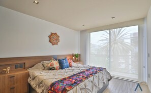 Una camera da letto con un letto, un comodino, un elemento decorativo a parete e vista su palme attraverso una porta scorrevole.