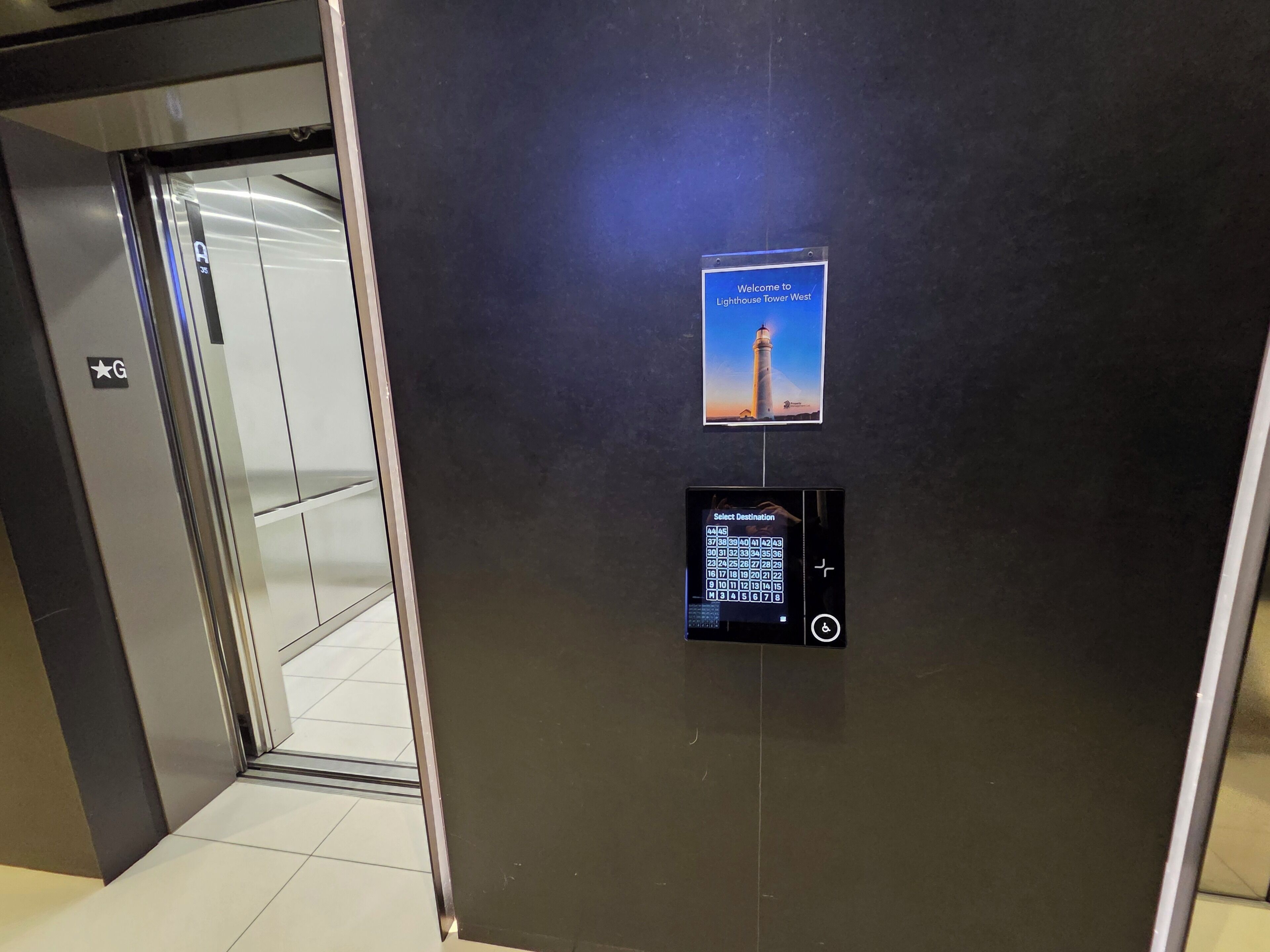 An elevator interior with a digital destination selector and a welcome sign for Lighthouse Tower West.