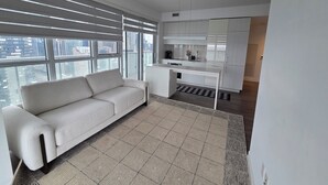 A modern living room with a sofa, a kitchen area with white cabinets, and large windows offering a city view.