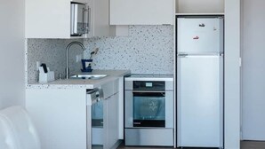 Una cocina compacta con gabinetes blancos, un refrigerador de acero inoxidable, un horno empotrado y una mesa de centro de vidrio.