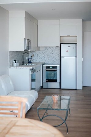 Eine kleine Küche mit weißen Schränken, einem Edelstahlkühlschrank, einem eingebauten Backofen und einem Glastisch.