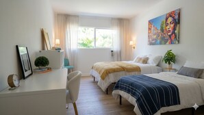 Et hotelværelse med et skrivebord, to senge, et billede og et vindue med gardiner.