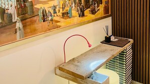 Eine Wand mit einem großen historischen Gemälde, das eine traditionelle Szene darstellt, eine hölzerne Bank mit Büchern und eine moderne Leuchte.