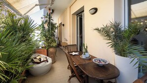 Ein Holztisch mit weißem Teller und einer Topfpflanze, eine hölzerne Bank und ein weißer Stuhl auf einem Balkon mit Blumentöpfen.