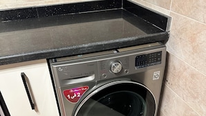 A compact washer and dryer unit with a control panel, located in a corner of a room with tiled flooring and a black countertop.