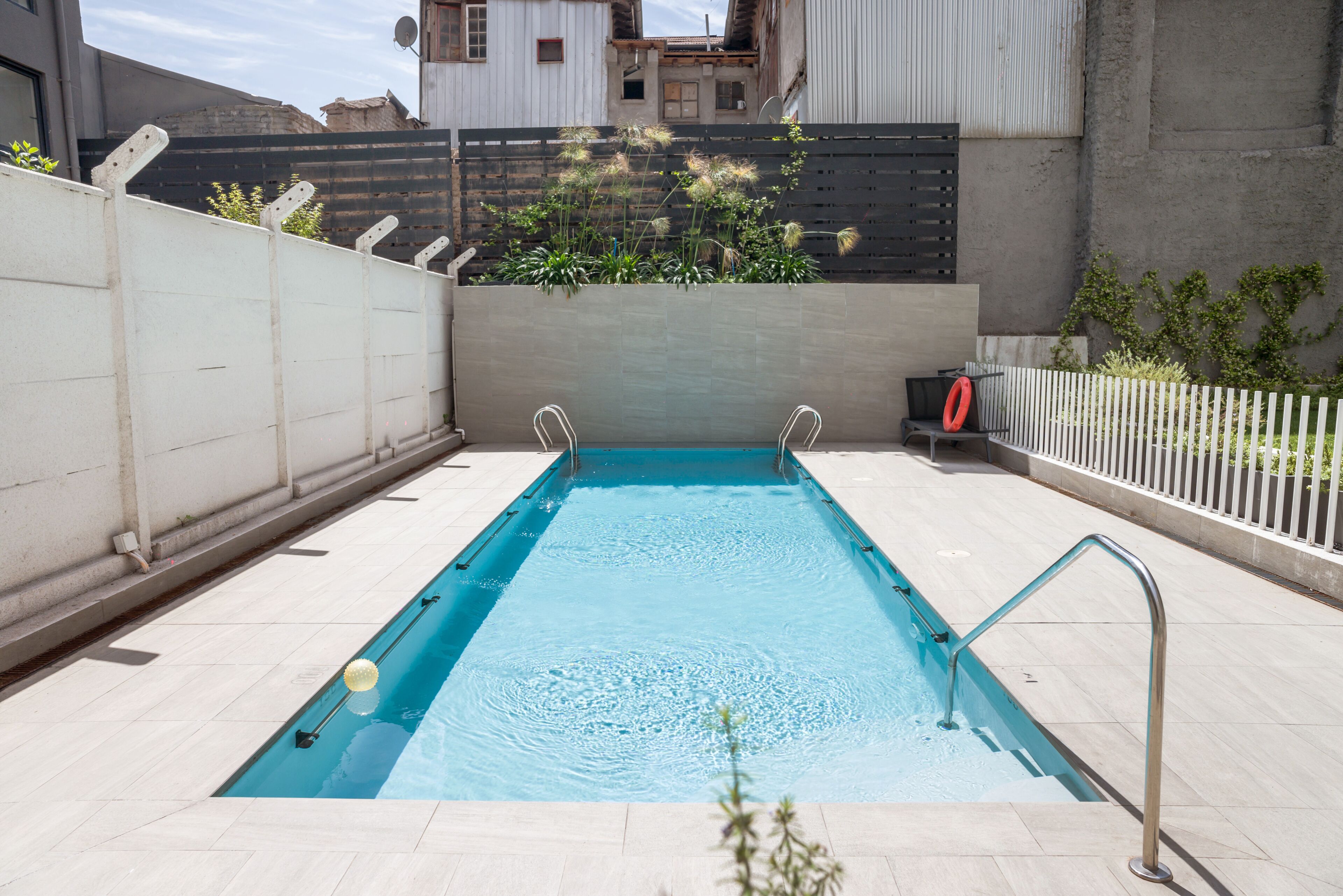 A rectangular swimming pool with a metal ladder on one side, surrounded by a white fence and a concrete wall.