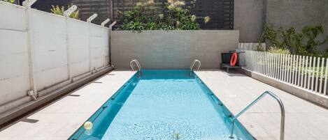 A rectangular swimming pool with a metal ladder on one side, surrounded by a white fence and a concrete wall.