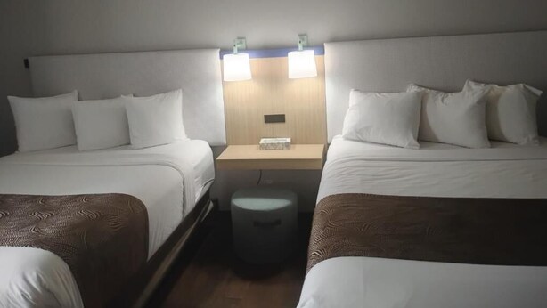 A hotel room with two beds, a nightstand, and a small stool. - Battle Creek Hotel (Battle Creek)