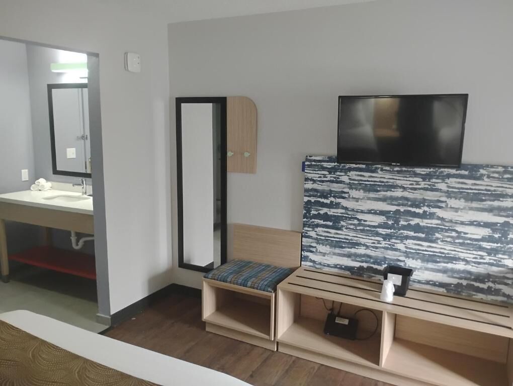 Habitación de hotel moderna con un banco de madera, un espejo grande, un televisor de pantalla plana y un baño con lavabo y espejo.