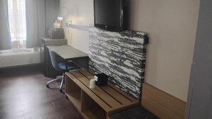 A hotel room with a TV mounted on a wall, a desk with a chair, a bench, and a window with curtains. - Battle Creek Hotel (Battle Creek)