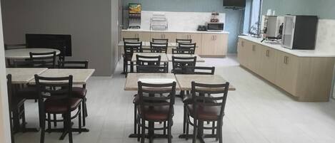A dining area with tables and chairs, a kitchen area with appliances, and a television mounted on the wall.