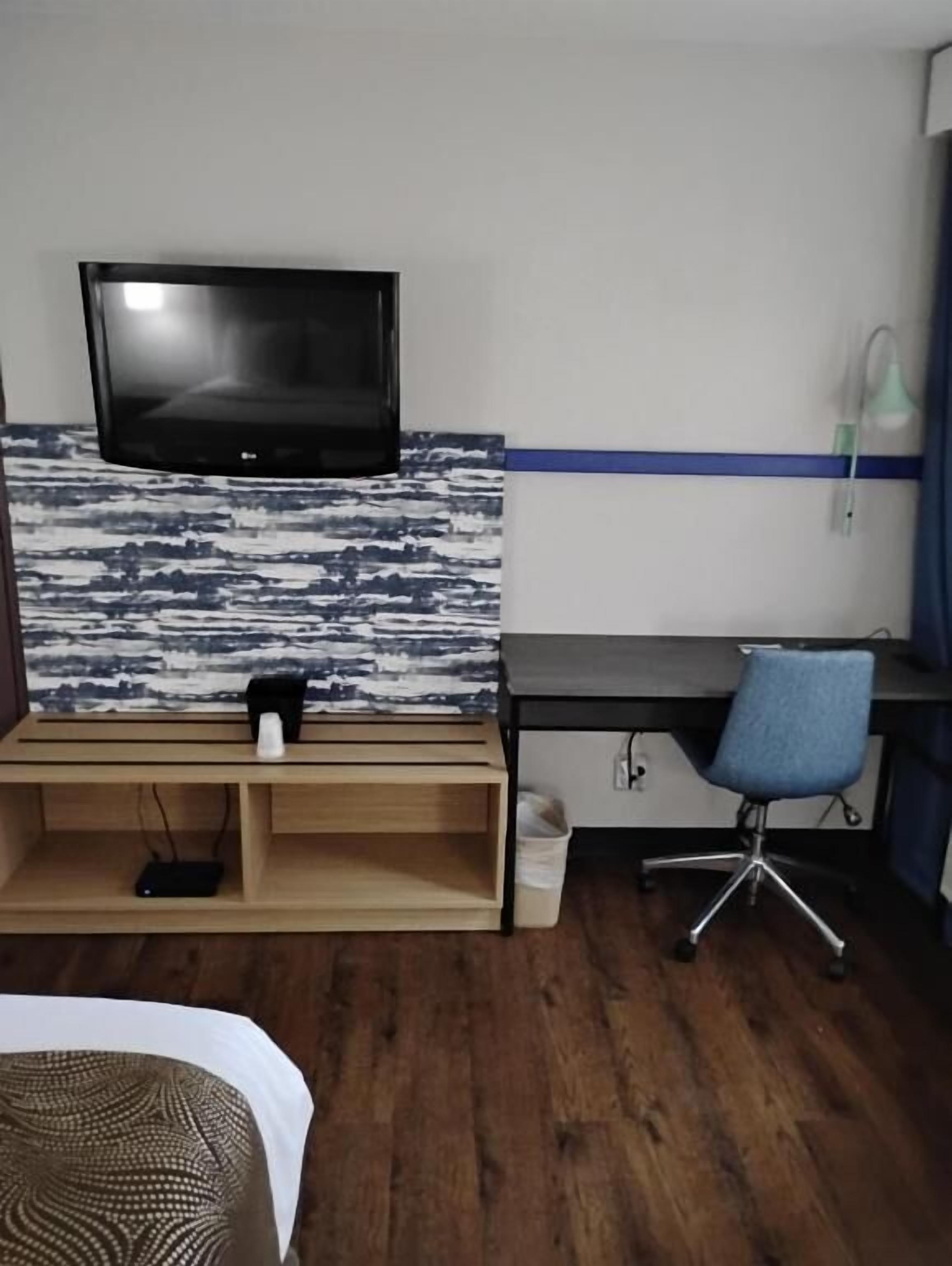 A room with a flat-screen TV mounted on a textured wall, a wooden desk with a chair, a lamp, and a waste bin.