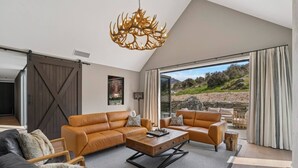 A spacious living room with a large window, a chandelier, and a fireplace. - Modern Alpine Retreat in Jacks Point (Queenstown)