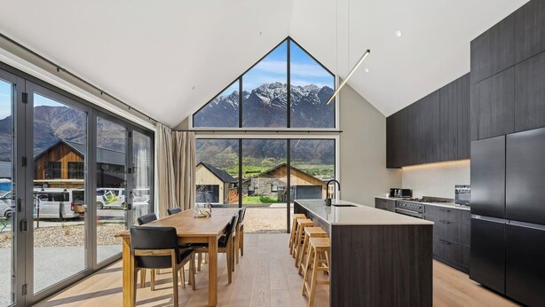 A modern kitchen with a large island, dark cabinetry, and a view of mountains through large windows. - Modern Alpine Retreat in Jacks Point (Queenstown)