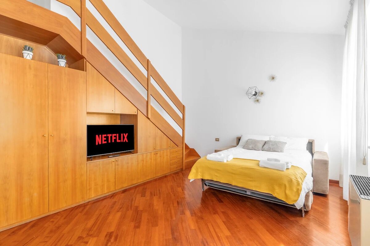 Ein Schlafzimmer mit einer Holztreppe, einem Bett mit gelber Decke und einem Fernseher, der 'NETFLIX' anzeigt.