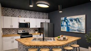 Uma cozinha com ilha central, armários brancos, eletrodomésticos de aço inoxidável e bancada com banquetas.