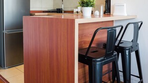 A modern kitchen with wooden cabinets, a stainless steel refrigerator, a microwave, a coffee maker, and a small island with two black bar stools. - 1BR in Heart of Providencia Urban Style (Providencia)