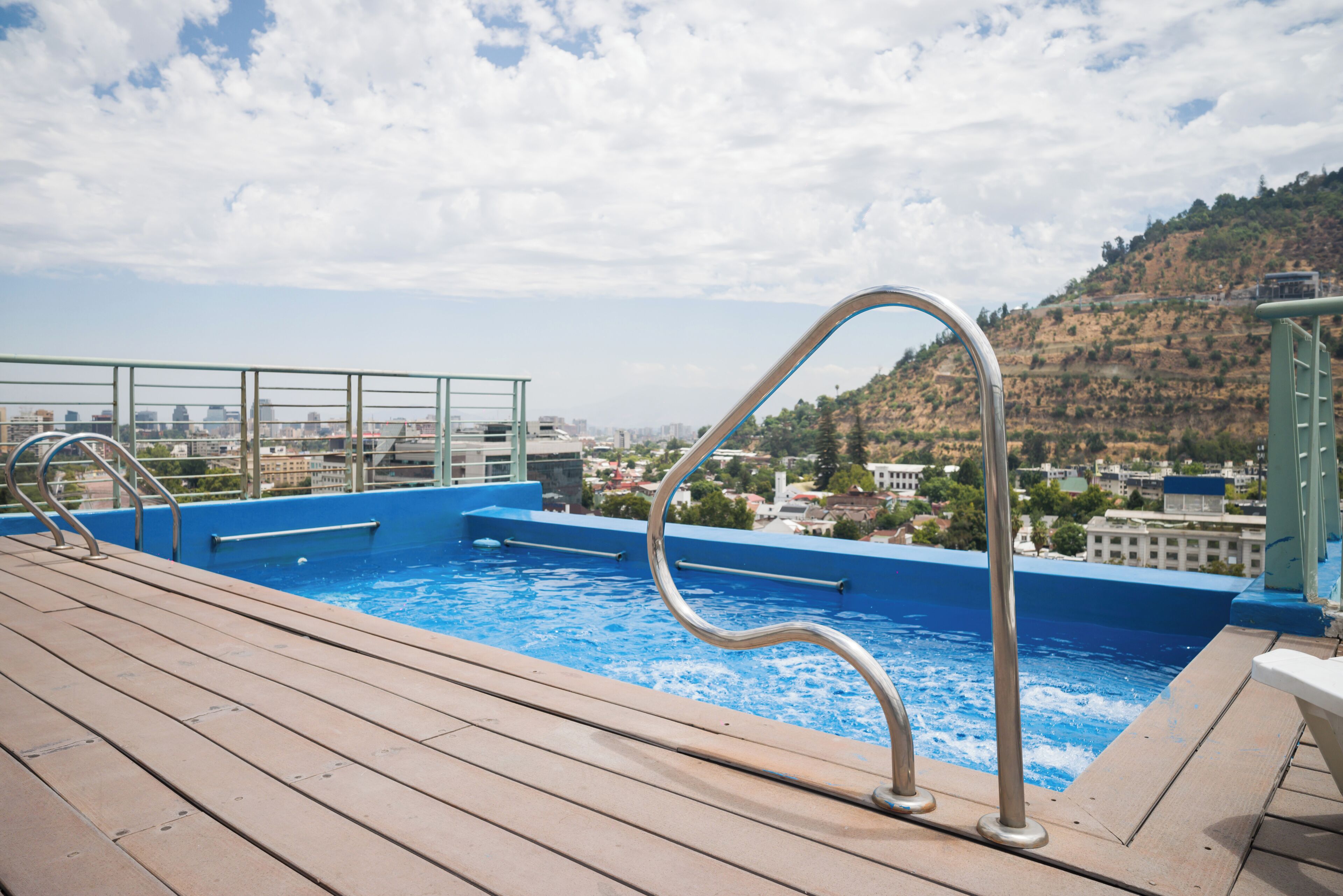 A swimming pool with a wooden deck and metal railings, overlooking a cityscape and hills.