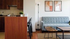 A modern kitchen with wooden cabinets, a dining area with a table and chairs, a grey sofa, and two framed artworks on the wall. - 1BR in Heart of Providencia Urban Style (Providencia)