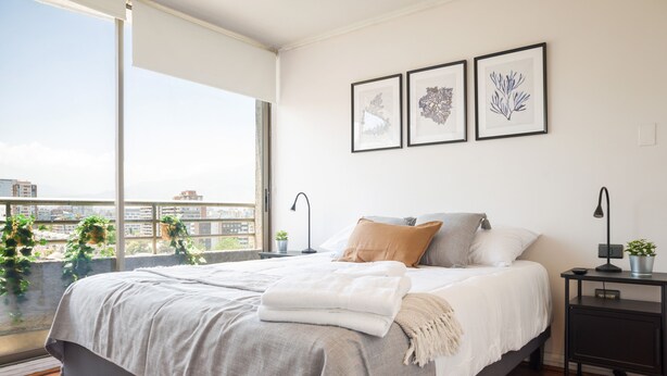 A bedroom with a large bed, a nightstand, a lamp, and a window with a city view. - 1BR in Heart of Providencia Urban Style (Providencia)